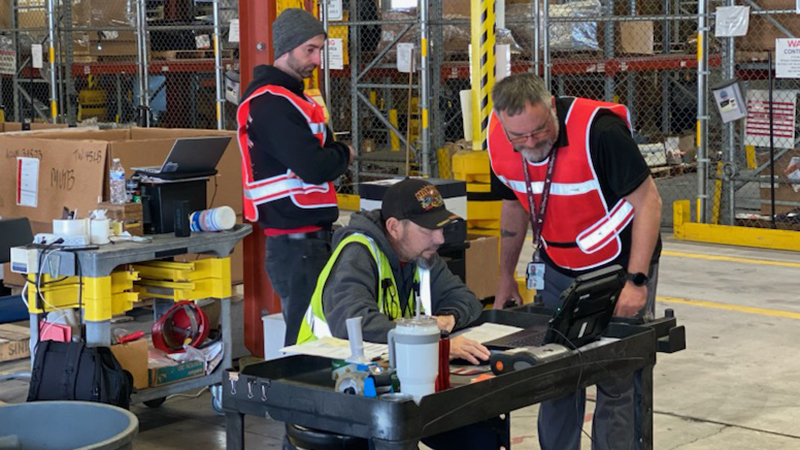 Three warehouse workers in safety gear using a computer in the middle of a warehouse